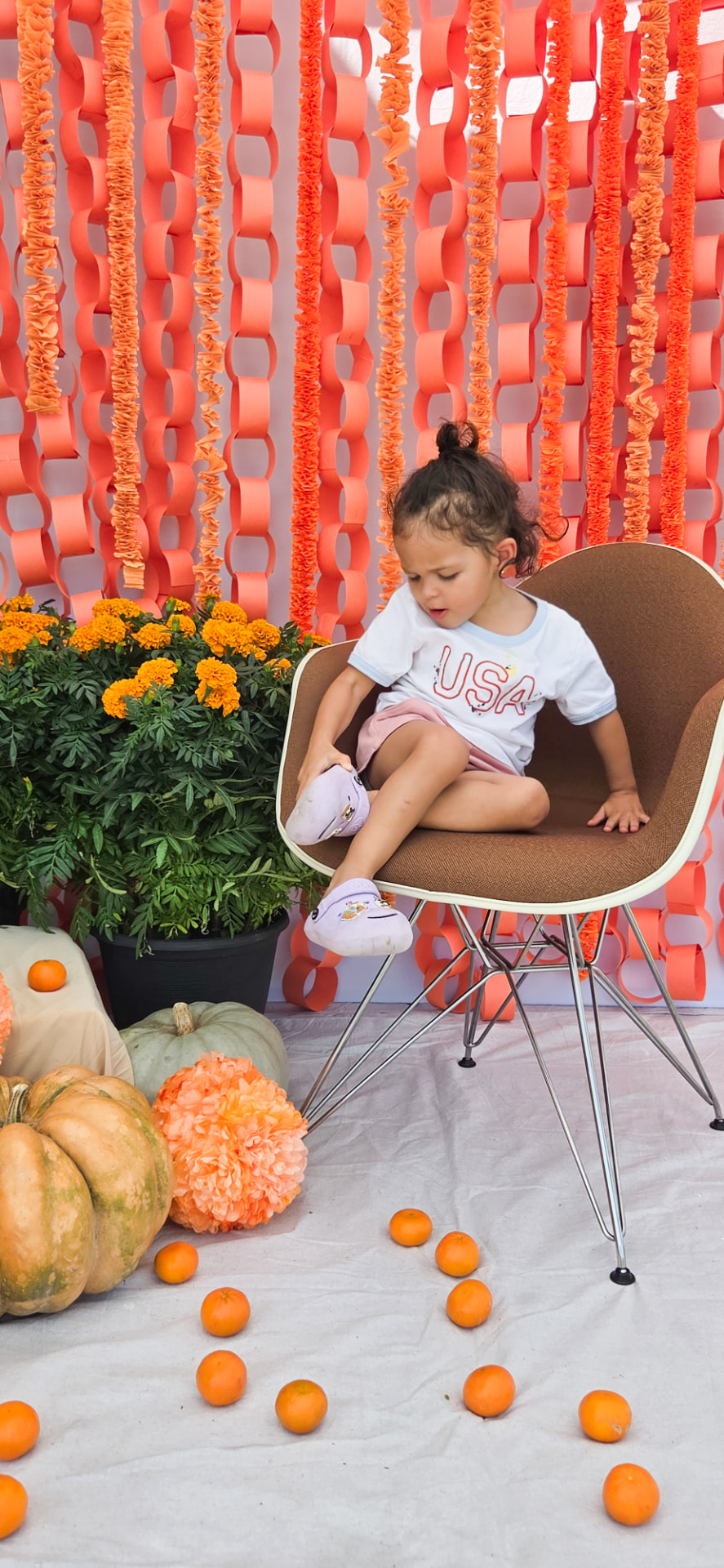 A child seated inside The Fall Paper Garden installation, surrounded by handmade orange paper chains, marigolds, and pumpkins